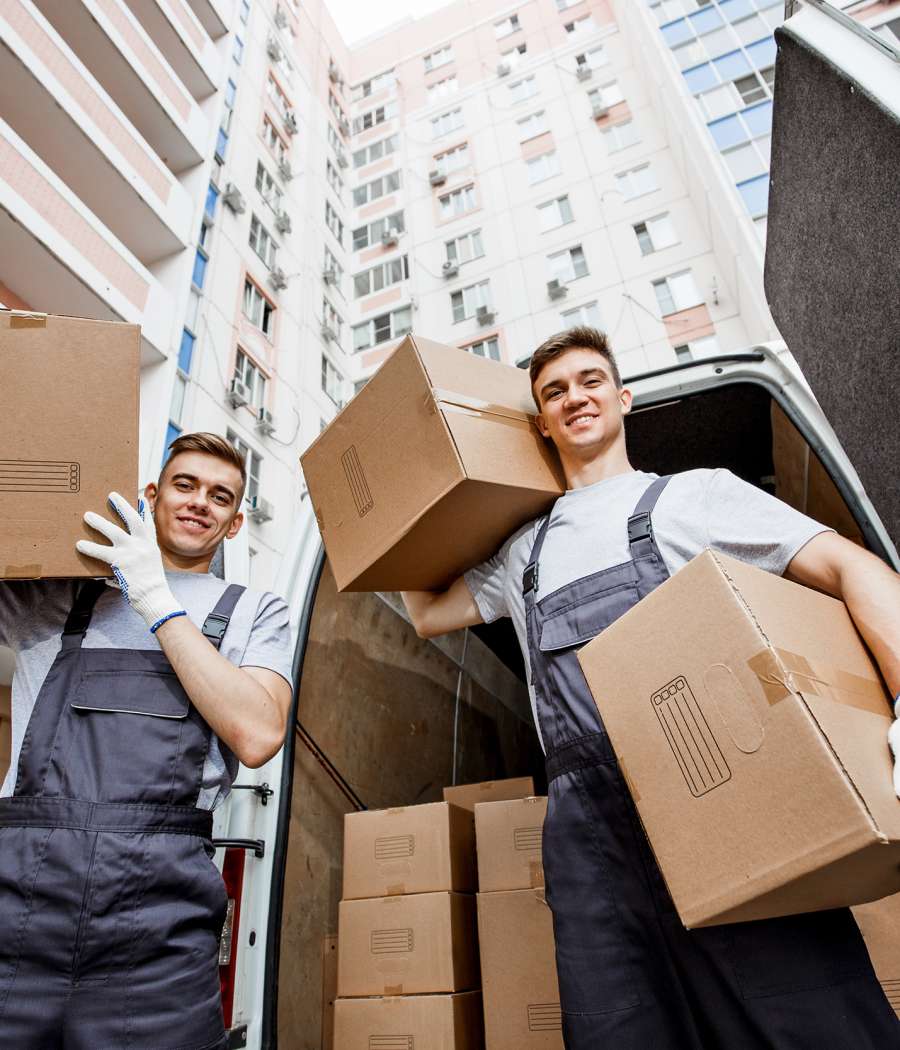 Two young handsome smiling workers wearing uniforms are standing in front of the van full of boxes holding boxes in their hands. The block of flats is in the background. House move, mover service.